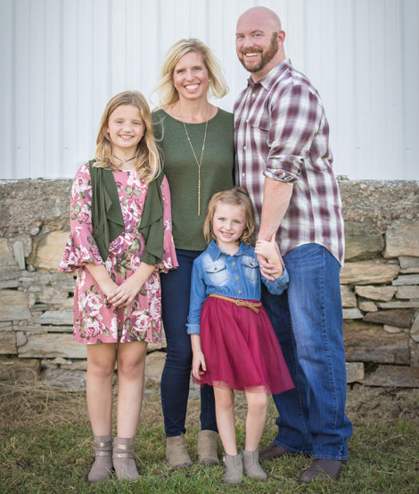 Photo showing Bill Healy Junior standing outside in front of a stone fence with his wife and two daughters